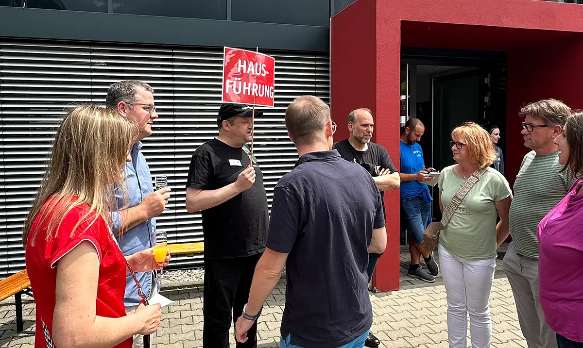 Gruppe an Menschen steht vor einem modernen Gebäude mit rotem Eingang. Eine Person hält ein Schild Hausführung.