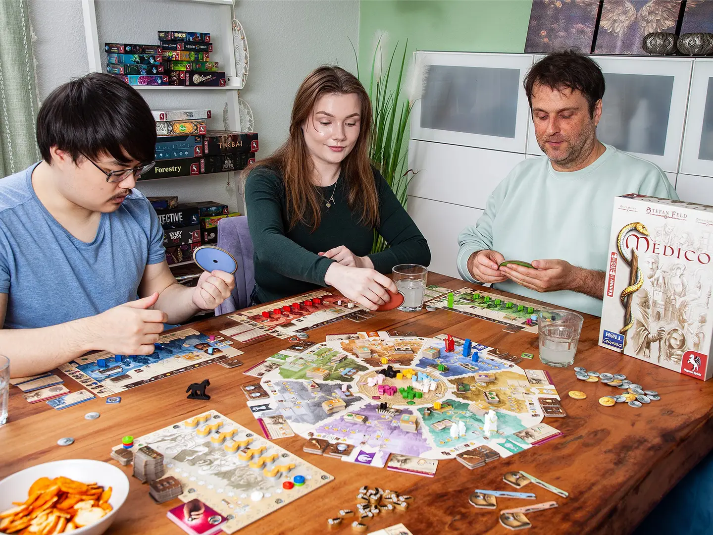 Drei Personen spielen gemeinsam Medico an einem Holztisch mit großem Spielplan, Spielertableaus, Karten und bunten Holzfiguren. Rechts steht die Spielschachtel.
