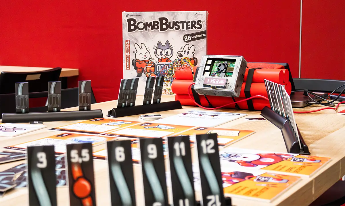 Das aufgebaute Brettspiel Bomb Busters mit Karten, Zahlenmarkern und einer Attrappe aus Dynamitstangen liegt auf einem Tisch vor rotem Hintergrund.