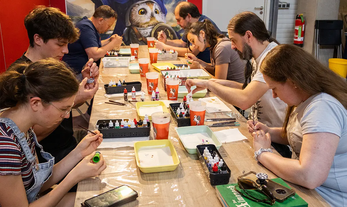 Gruppe an Menschen sitzt an einem Biertisch mit Bechern, Farben und Pinseln und bemalt Miniaturen.