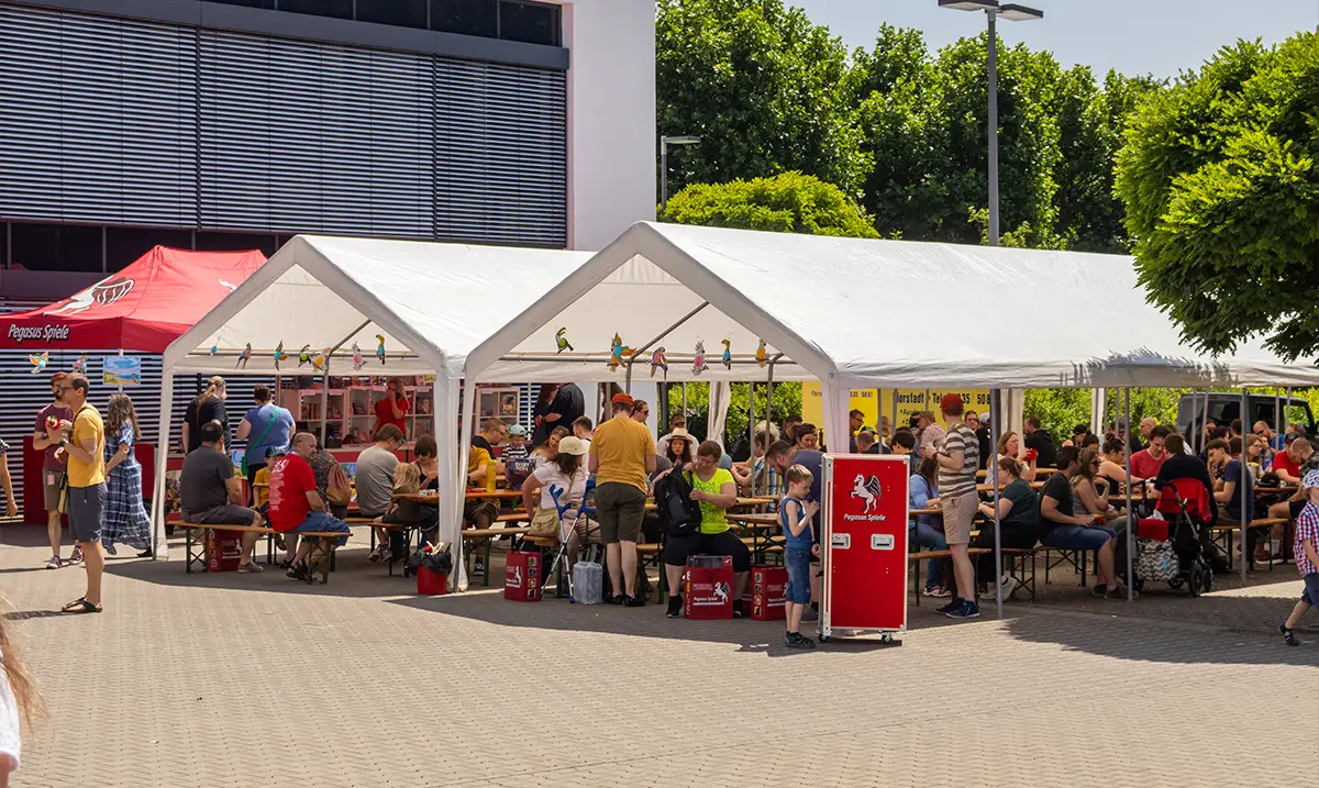 Menschen sitzen und stehen an langen Bierbänken unter mehreren weißen Festzelten auf einem sonnigen Außengelände. Im Hintergrund sind Bäume und ein modernes Gebäude zu sehen.