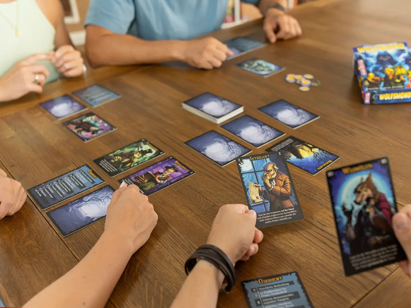 Nahaufnahme einer Spielrunde Wolfsmond mit ausgelegten Karten auf einem Holztisch. Im Vordergrund hält eine Person die Karte Intrigant, eine weitere Person hält eine Karte mit einer Wolfsgestalt.