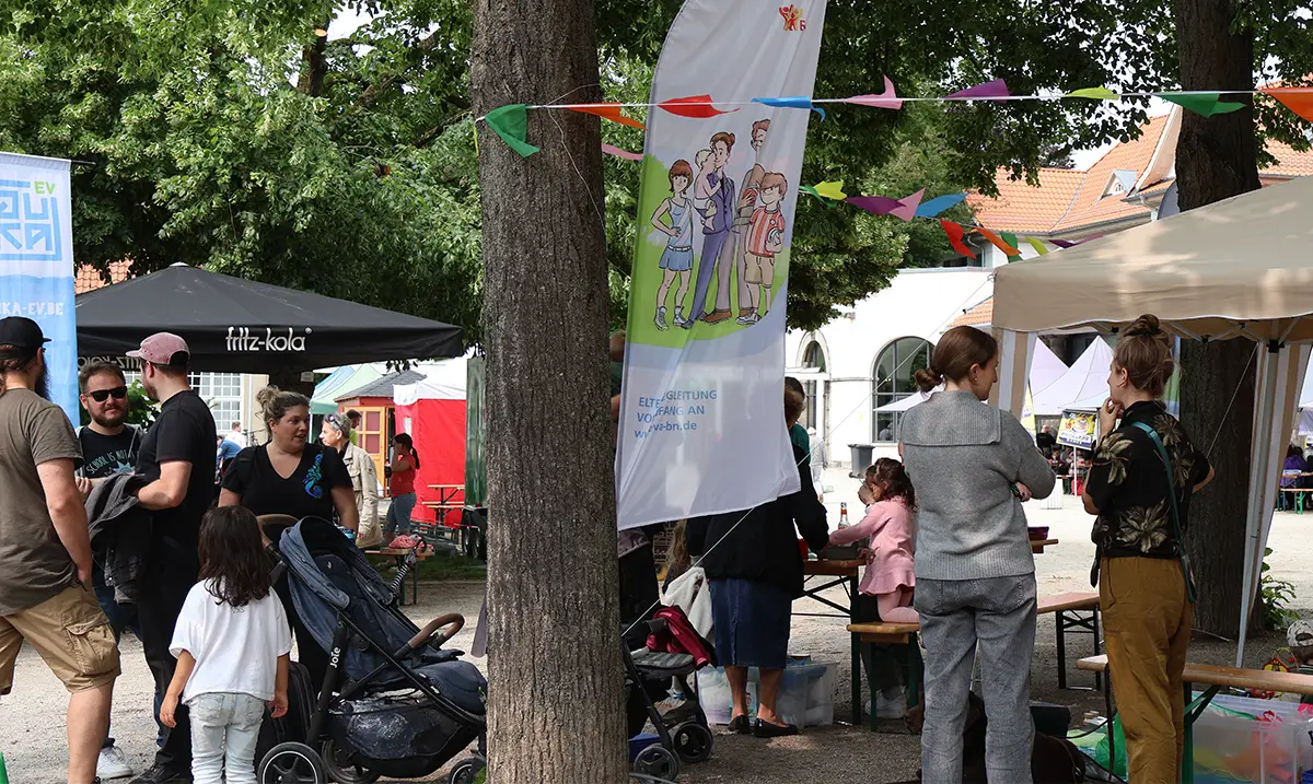 Personen stehen um Biertische und Bänke neben einem Baum