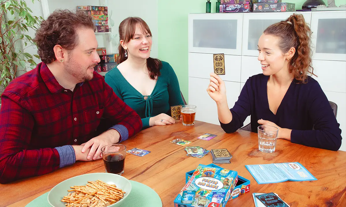 Drei Personen sitzen am Tisch und spielen das Kartenspiel FERTIG ICH HABE