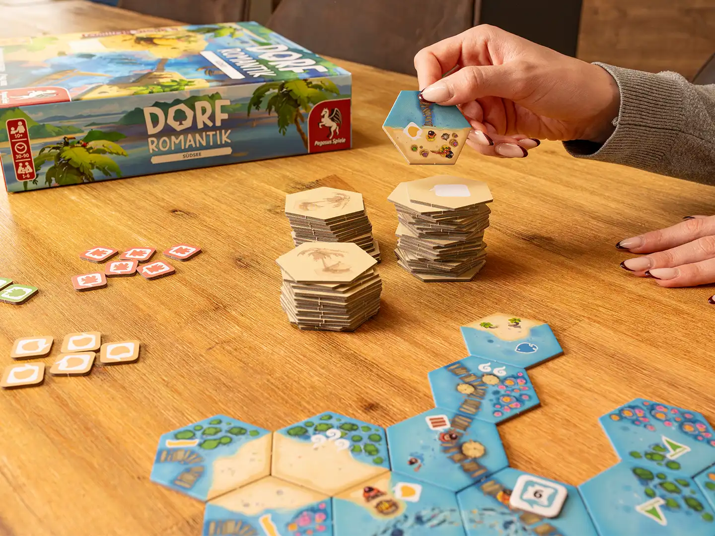 Eine Person hält ein sechseckiges Landschaftsplättchen über einem Holztisch mit ausgelegten Insel- und Meeresplättchen, Spielmarkern und Plättchenstapeln aus dem Brettspiel Dorfromantik – Südsee. Im Hintergrund steht die Spielschachtel.