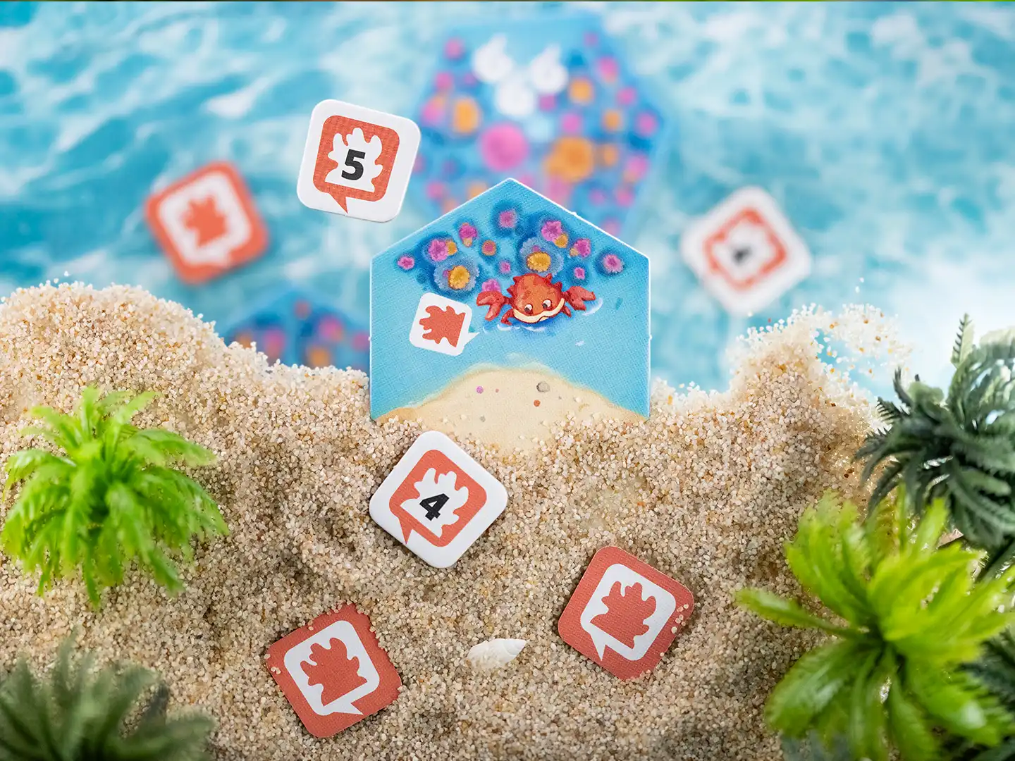 Sechseckiges Spielplättchen mit roter Krabbe und bunten Korallen aus dem Brettspiel Dorfromantik – Südsee, umgeben von Spielmarkern mit Krabbensymbolen und Zahlen auf Sand zwischen grünen Pflanzen