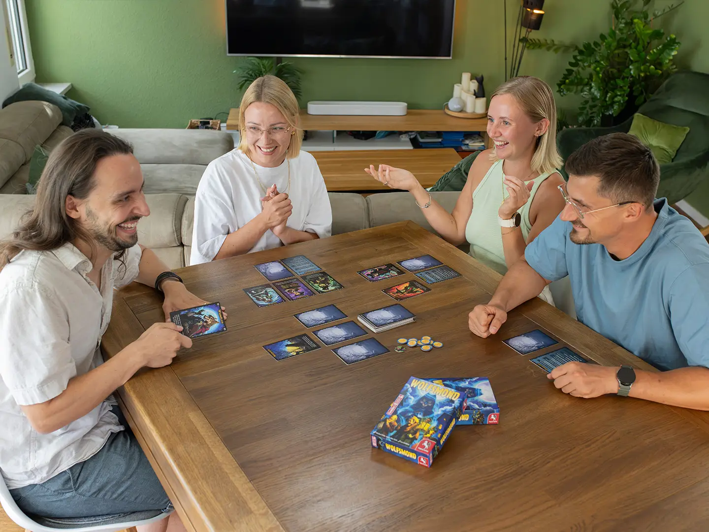 Vier Personen lachen gemeinsam beim Kartenspiel Wolfsmond. Auf dem Holztisch liegen Spielkarten, Marker und die Spielschachtel.